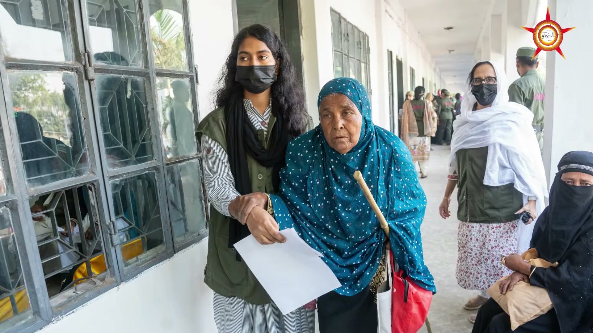 A younger woman walking beside an older woman at the medical camp; both hold papers.