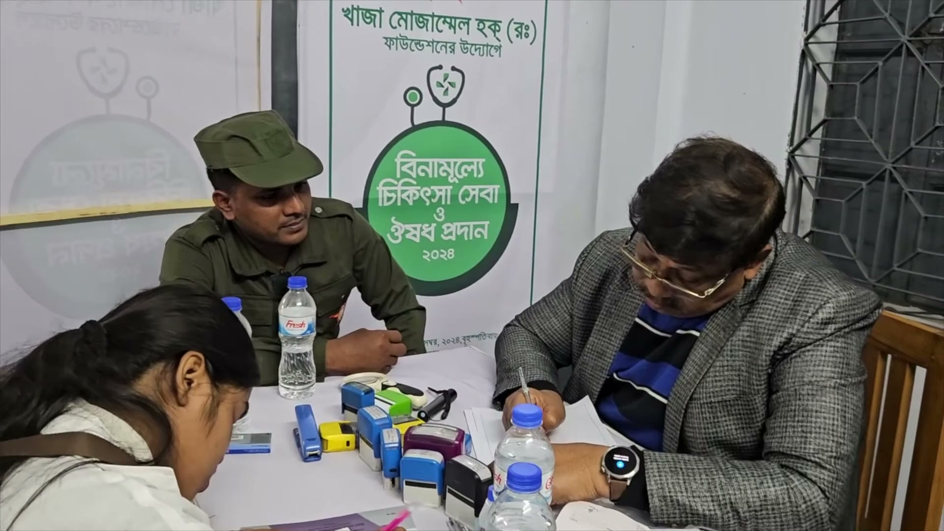 People at a consultation table with medicines, beside a KMRF camp sign.