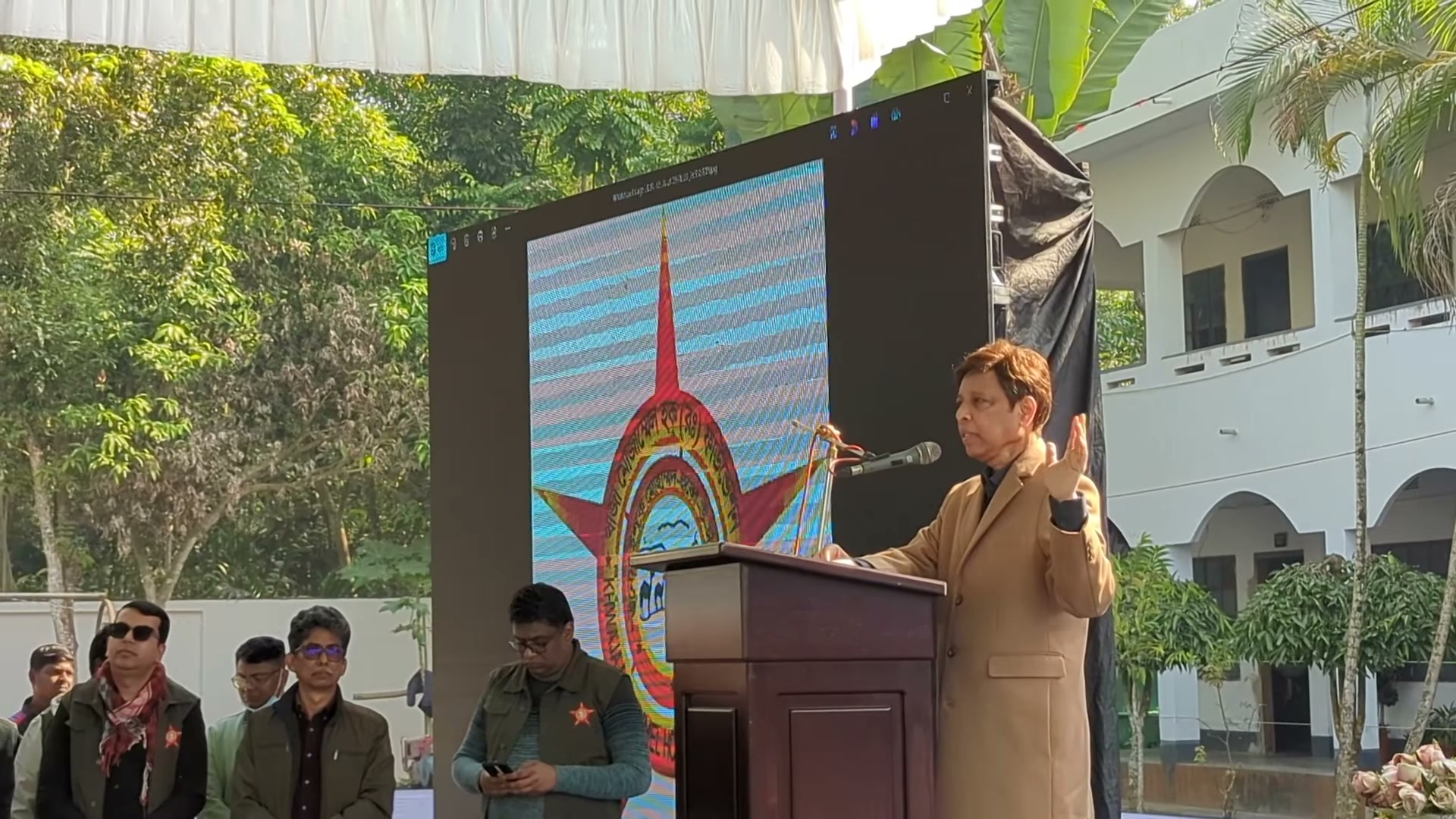The named speaker addressing the camp from a lectern, with the KMRF emblem on a screen behind.